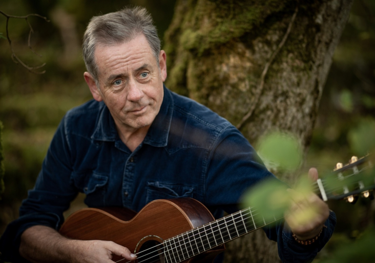 Irish singer-songwriter Luka Bloom holds an acoustic guitar while sitting outdoors against a mossy tree trunk in a natural forest setting.